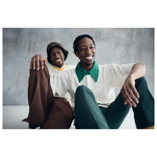 Two smiling young Black men pose indoors, wearing casual vintage-style shirts and slacks, with one arm of each resting on the other.