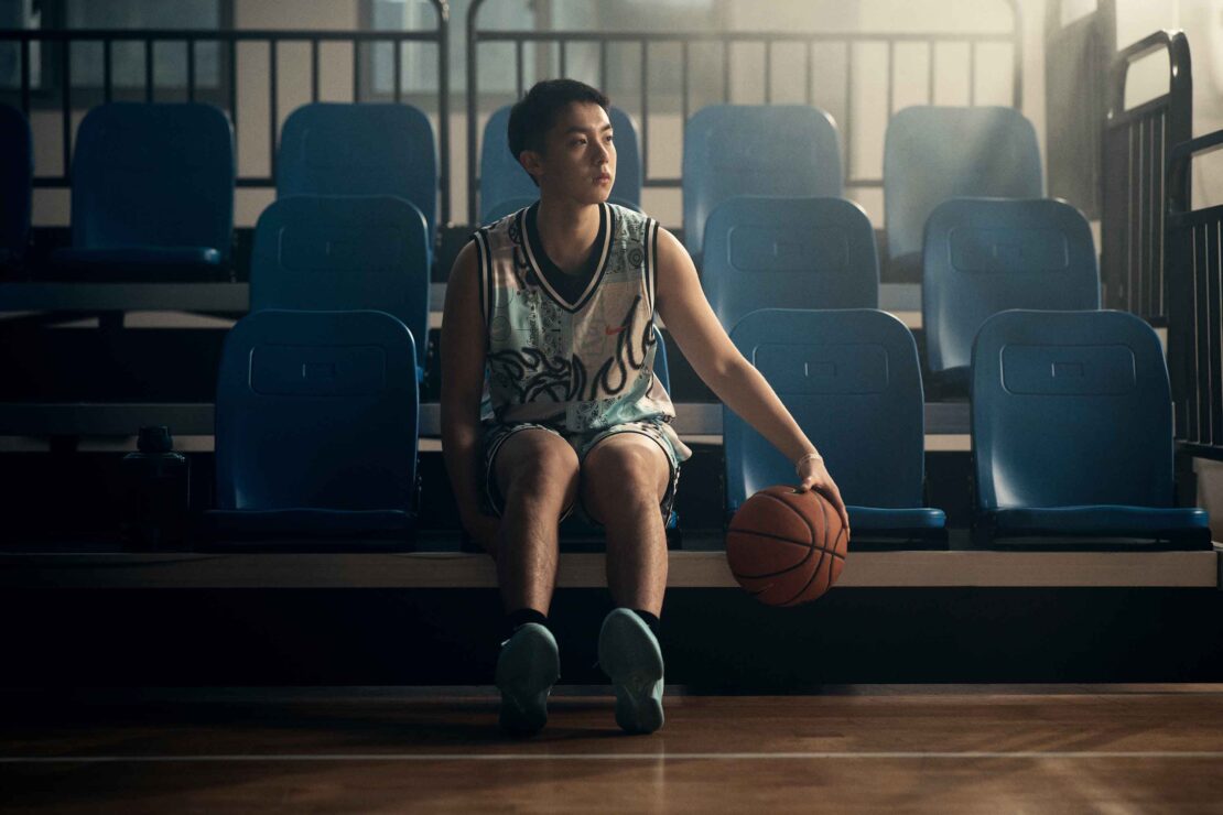 Young man in basketball jersey seated on bleachers holding a basketball by Olly Burn