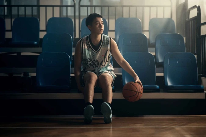 Young man in basketball jersey seated on bleachers holding a basketball by Olly Burn
