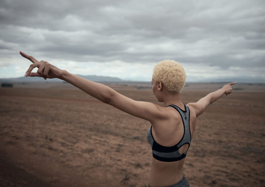 Athletic woman in sports bra stands in a field with arms outstretched, pointing. Cloudy sky backdrop, wellness and outdoor fitness concept.