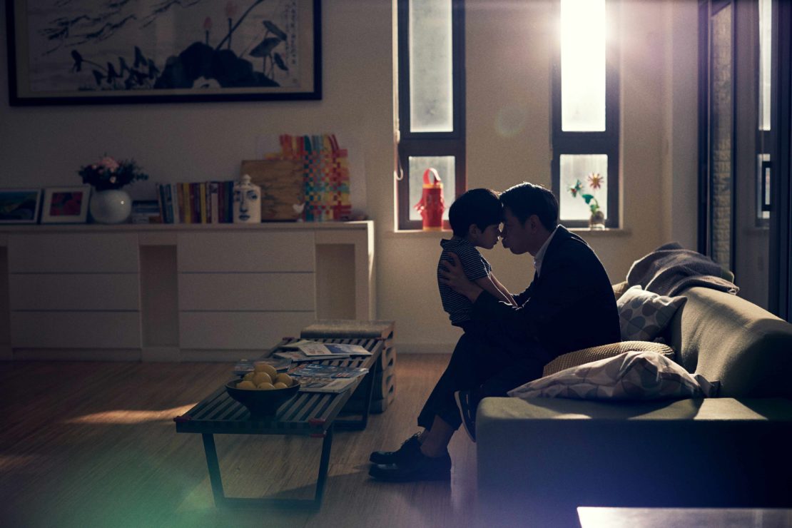 Indoor scene captures a father and son embracing in a warm living room, bathed in soft light from nearby windows, creating an intimate moment.