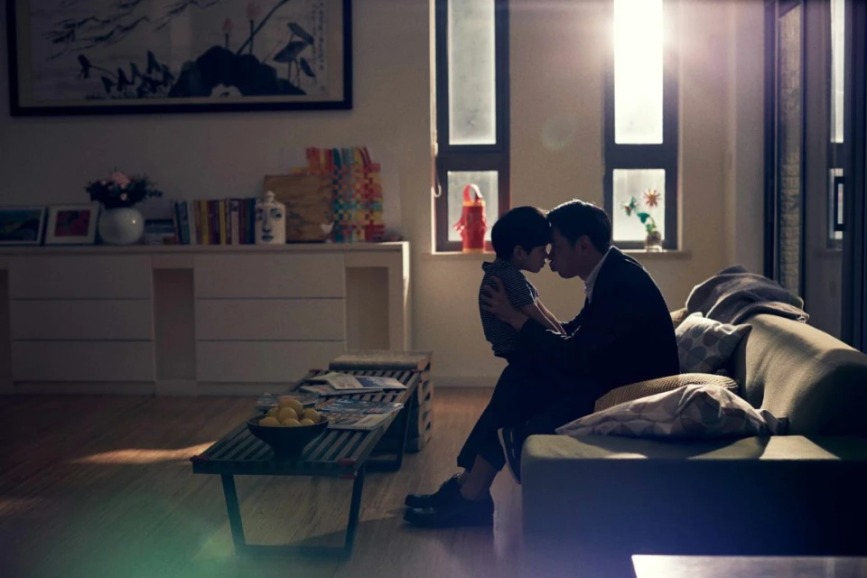 Indoor scene captures a father and son embracing in a warm living room, bathed in soft light from nearby windows, creating an intimate moment.
