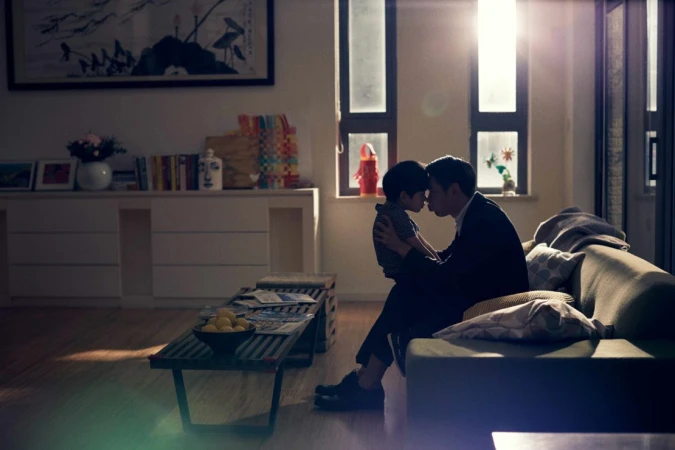 Indoor scene captures a father and son embracing in a warm living room, bathed in soft light from nearby windows, creating an intimate moment.