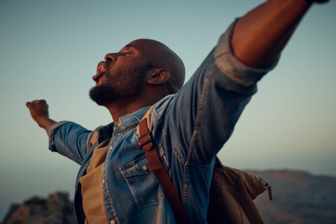 African American man stands on mountain with arms raised against sky, wearing denim jacket and backpack. Freedom and success concept.