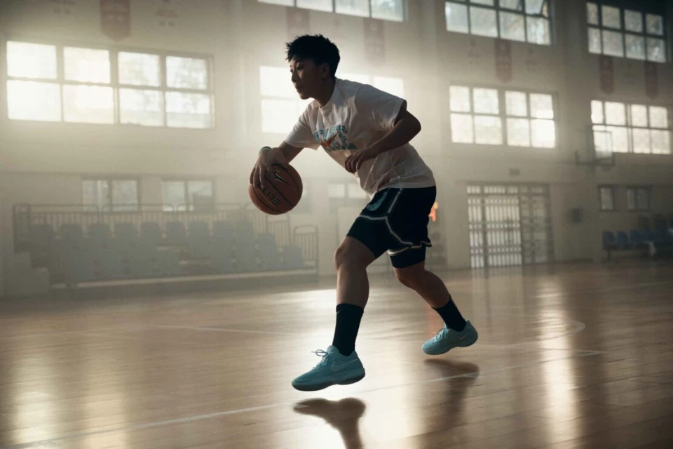 A young adult male in athletic wear dribbles a basketball on a polished indoor court, illuminated by soft, diffused light and haze.