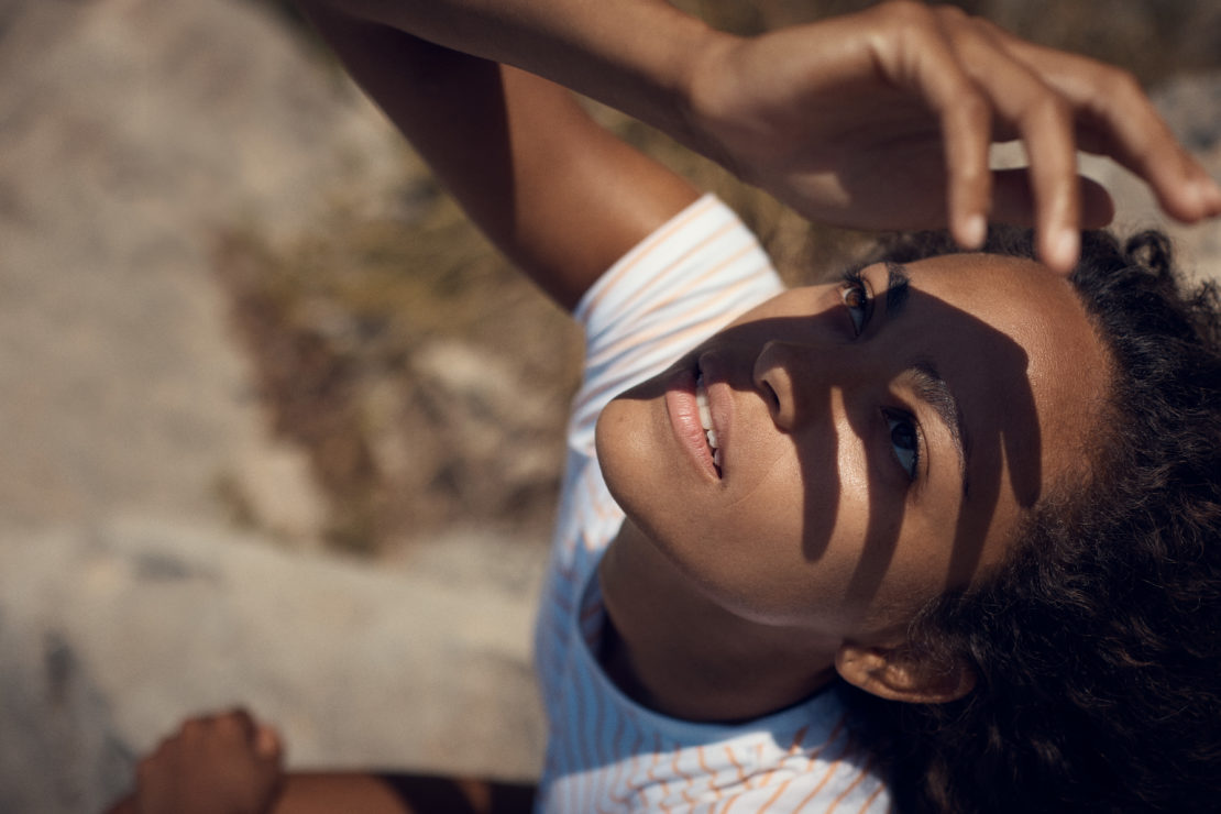 Close-up portrait of a Black woman with hand shadows on her face, wearing a striped top, posing outdoors in natural sunlight.
