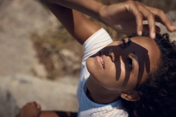 Close-up portrait of a Black woman with hand shadows on her face, wearing a striped top, posing outdoors in natural sunlight.