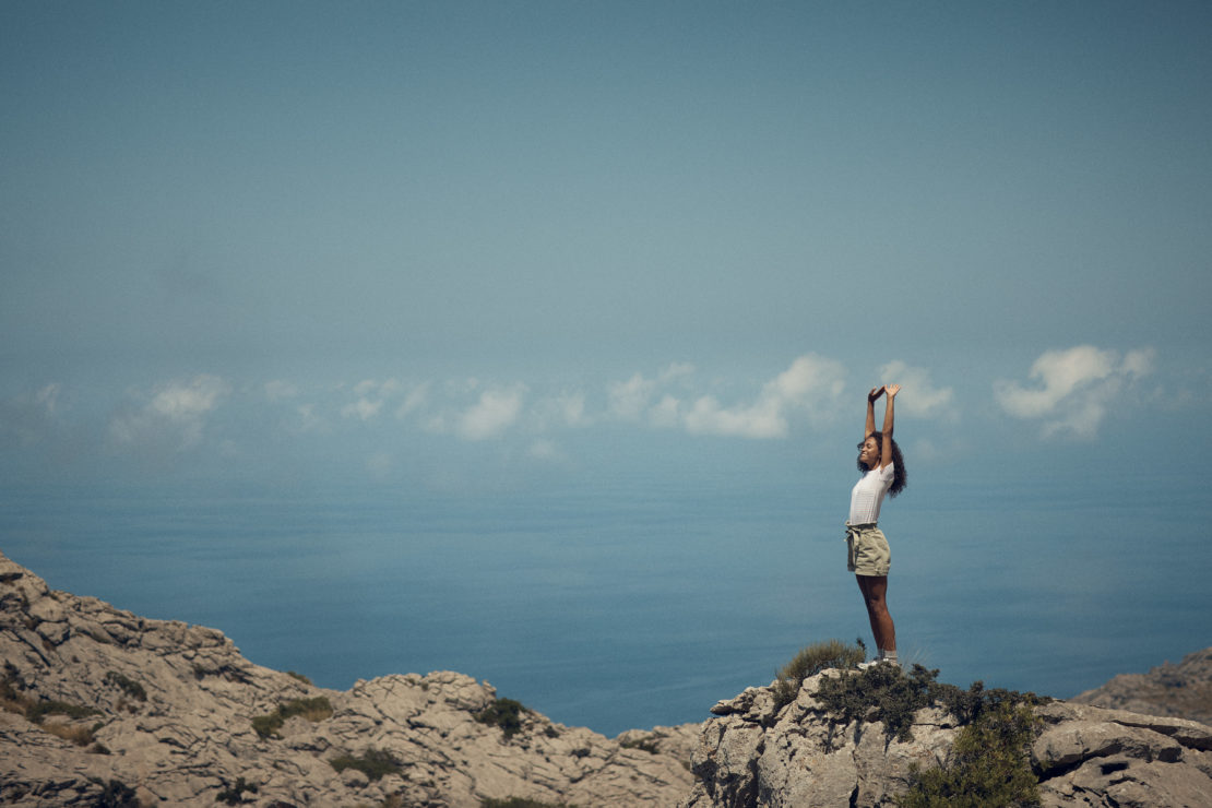 A woman standing on a rocky mountaintop with arms raised against a backdrop of blue sky and ocean, enjoying the scenic vista.