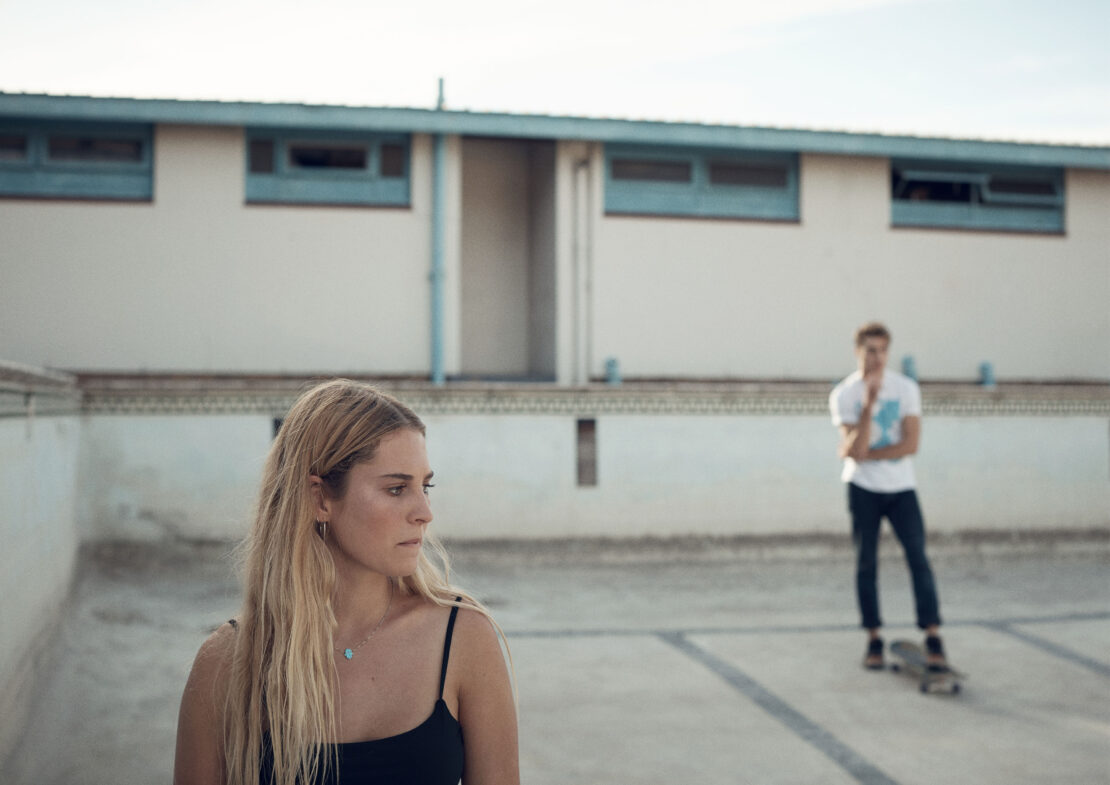 A woman and a man standing in an empty swimming pool with a white building in the background, wearing casual clothing.