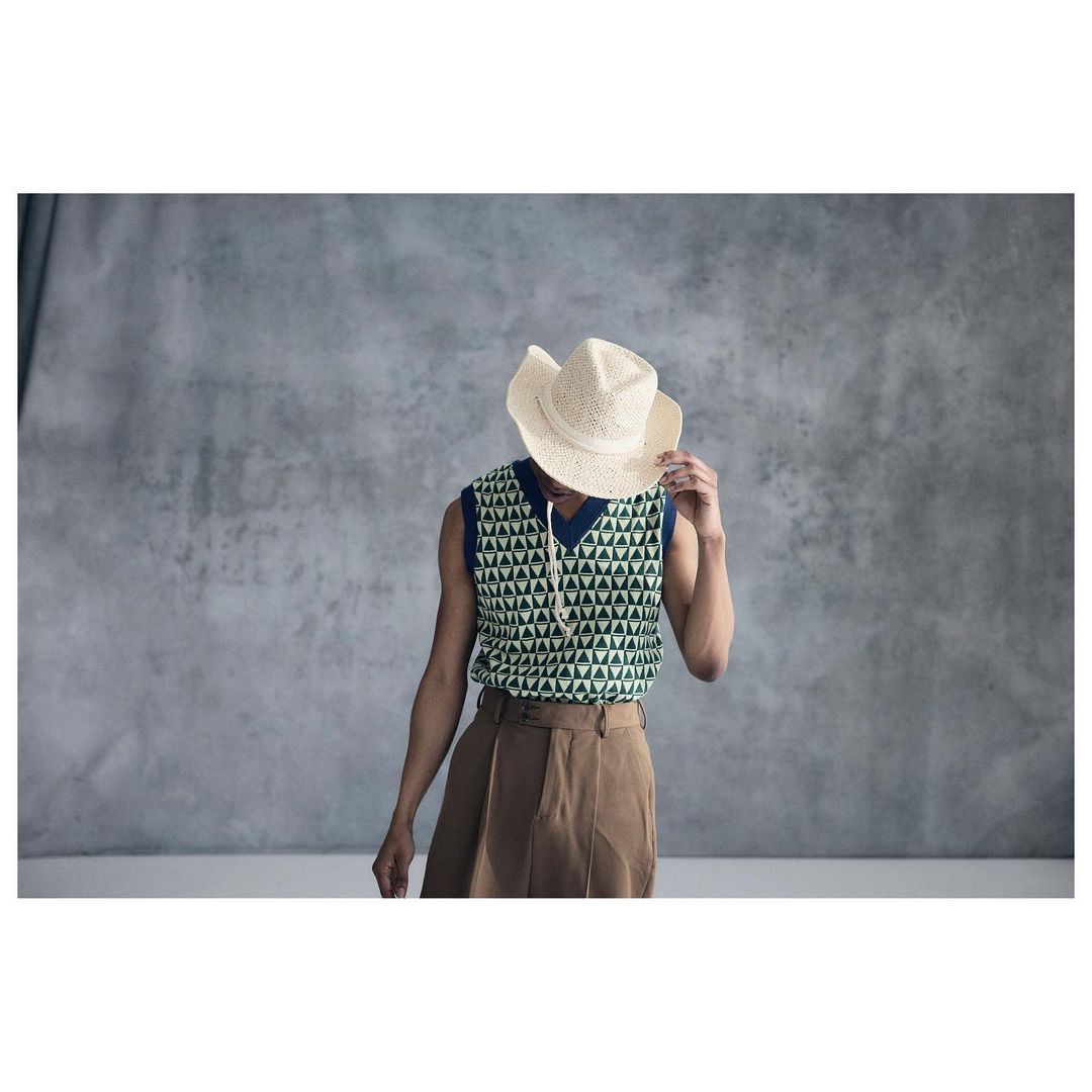 Male model poses in a patterned vest, tan pants, and a cowboy hat against a gray backdrop. Fashion editorial style.