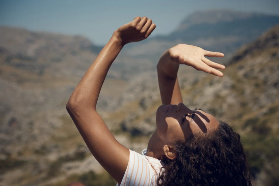 Woman outdoors in sunlight shields eyes with her hand, mountain background, relaxed expression, wearing striped shirt, clear blue sky.