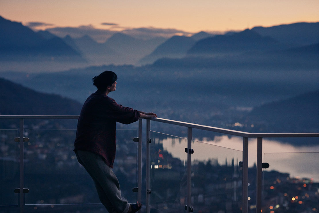 Adult female stands on a balcony overlooking a mountain range at twilight, city lights visible in the distance and clear railing in view.