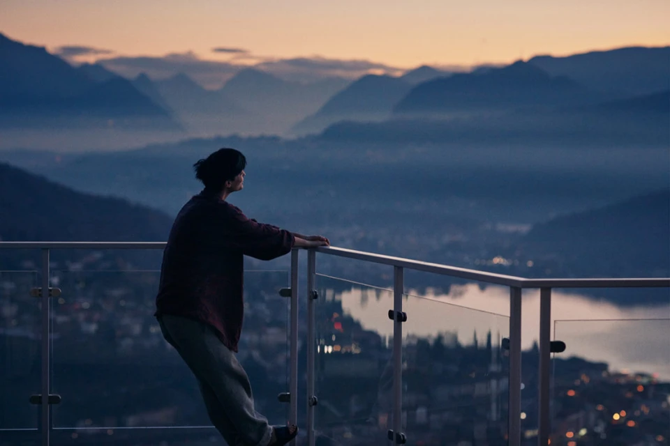 Adult female stands on a balcony overlooking a mountain range at twilight, city lights visible in the distance and clear railing in view.