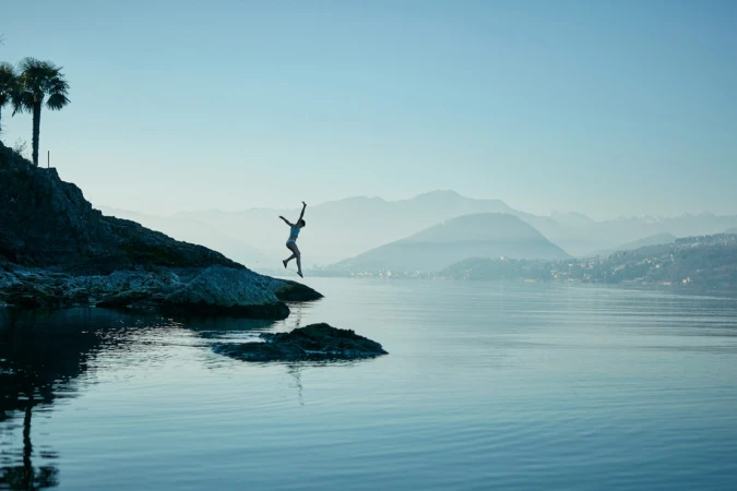 A person diving from a rocky cliff into a wide lake with mountains in the background under a pale sky.