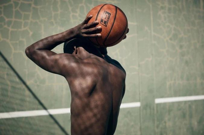 Shirtless black man holding a basketball behind his head against a green weathered court wall