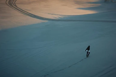 Aerial view of a person walking across vast sand dunes with tire tracks under golden hour light