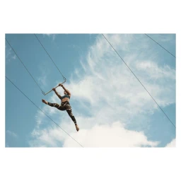 A female acrobat performing mid-air on a trapeze bar against a bright blue sky with wispy white clouds.