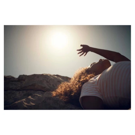 Person lying on rocky ground reaching up toward a bright sunlit sky in a low angle shot by Olly Burn
