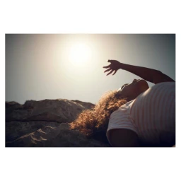 Person lying on rocky ground reaching up toward a bright sunlit sky in a low angle shot by Olly Burn