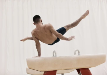 Male gymnast performing a one-arm handstand on a pommel horse against a pale curtained background