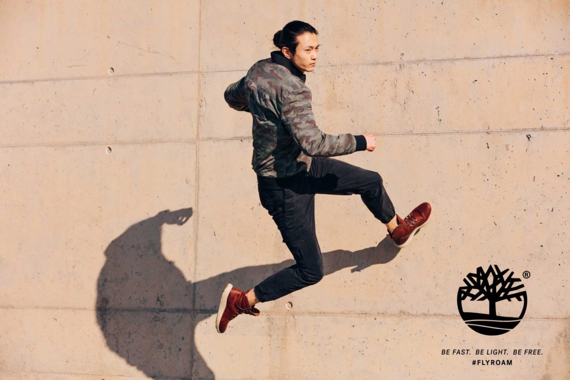Athletic man jumps mid-air in a fashion shot against a concrete wall, wearing sneakers and a camouflage jacket, showcasing movement and style.