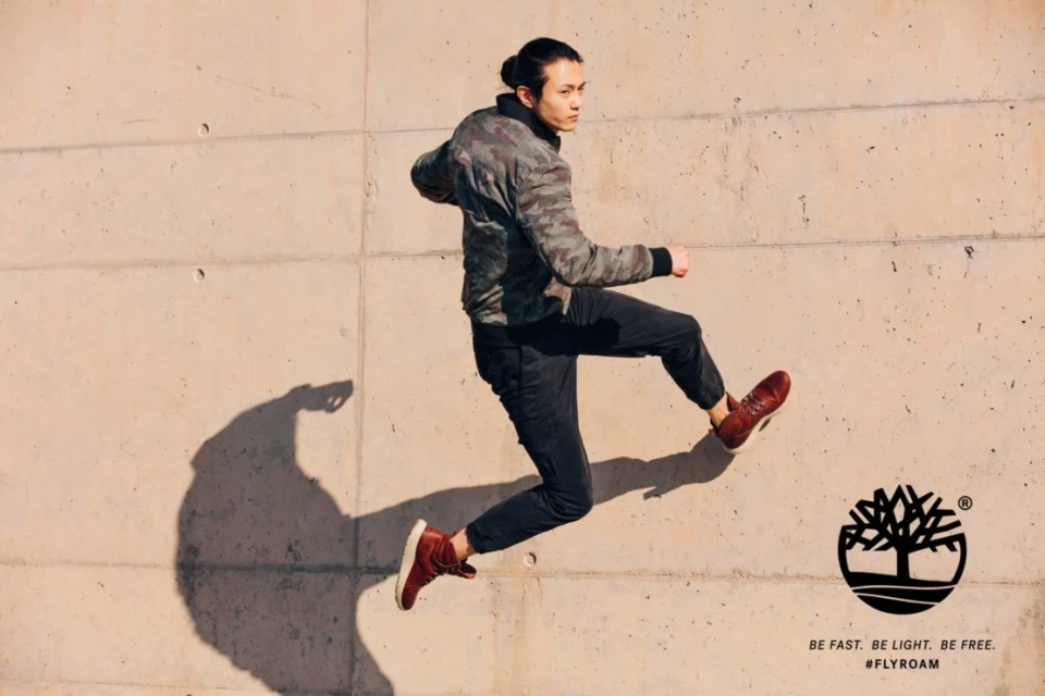 Athletic man jumps mid-air in a fashion shot against a concrete wall, wearing sneakers and a camouflage jacket, showcasing movement and style.