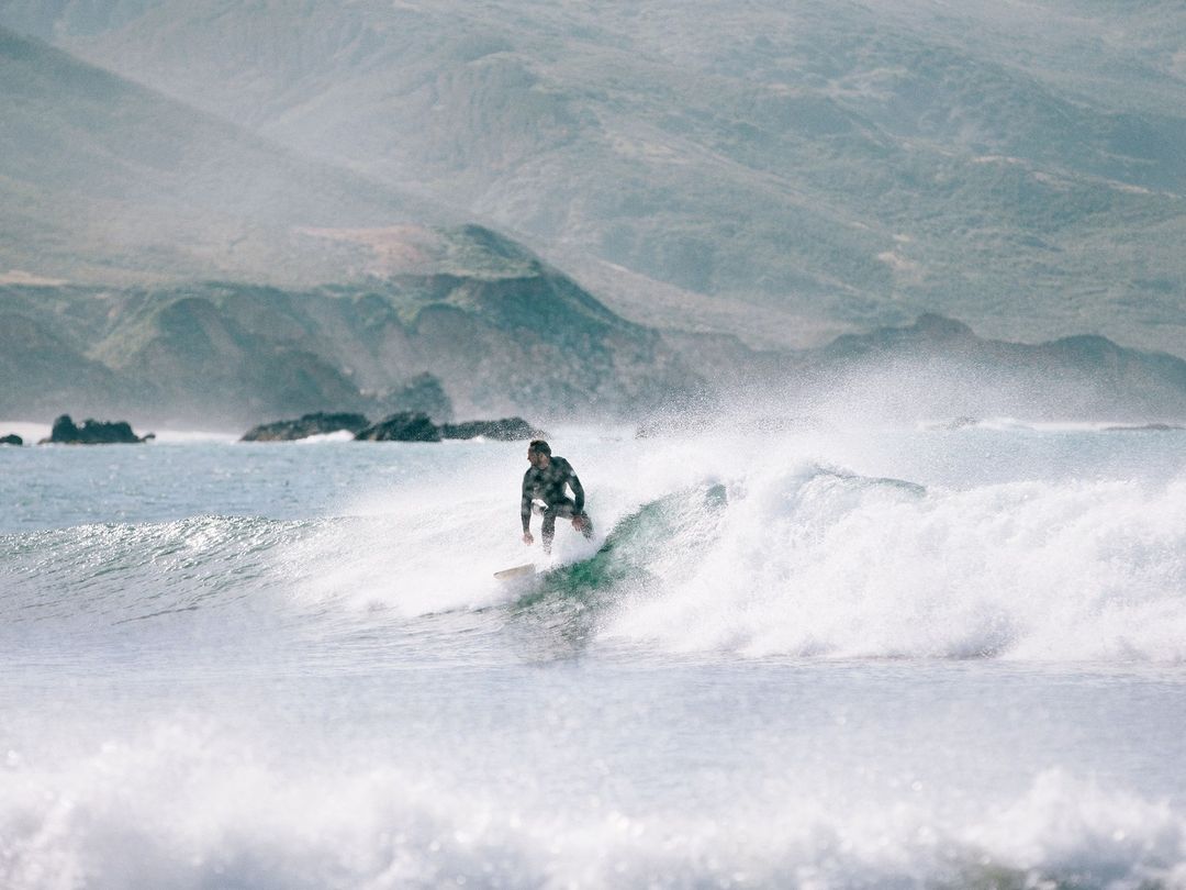 Surfer rides a wave on a surfboard in a wetsuit. Ocean with mountains in the distance under cloudy, overcast sky.