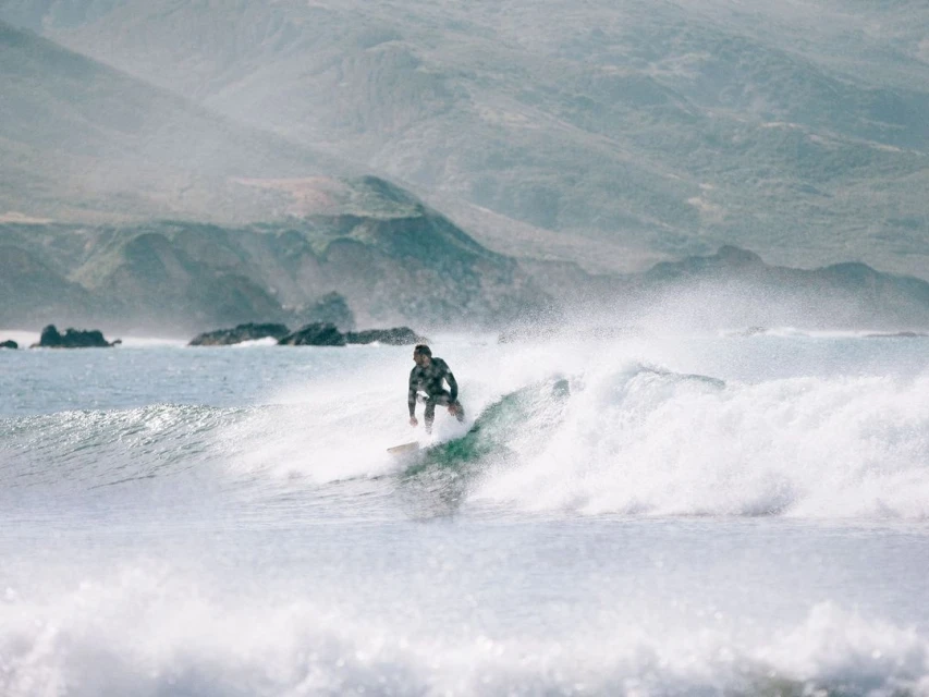 Surfer rides a wave on a surfboard in a wetsuit. Ocean with mountains in the distance under cloudy, overcast sky.