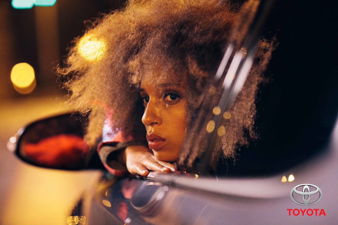Portrait of a young woman with curly hair, leaning on a car window at night. The Toyota logo is visible in the lower right corner.