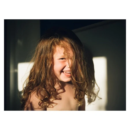 Portrait of a smiling child with long curly brown hair partially covering their face in natural light.