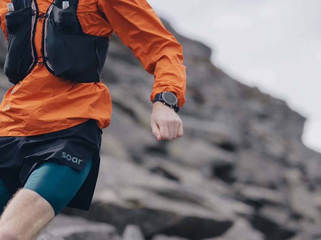 An athletic adult runs along a rocky mountain path, wearing a running vest, orange long-sleeved shirt, dark shorts, and a wristwatch.