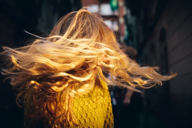 Woman with long blonde hair tossing her head, wearing a yellow sweater against an out-of-focus street background