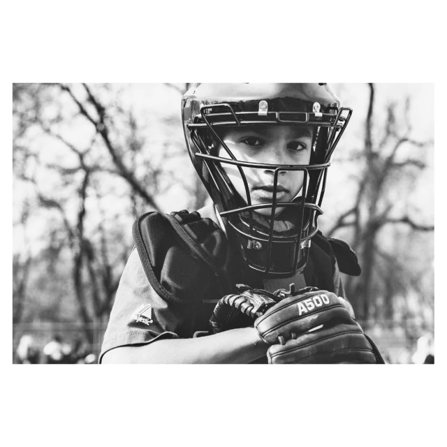 A monochrome portrait of a young baseball catcher wearing a helmet, chest protector, and mitt, with a blurred outdoor backdrop.
