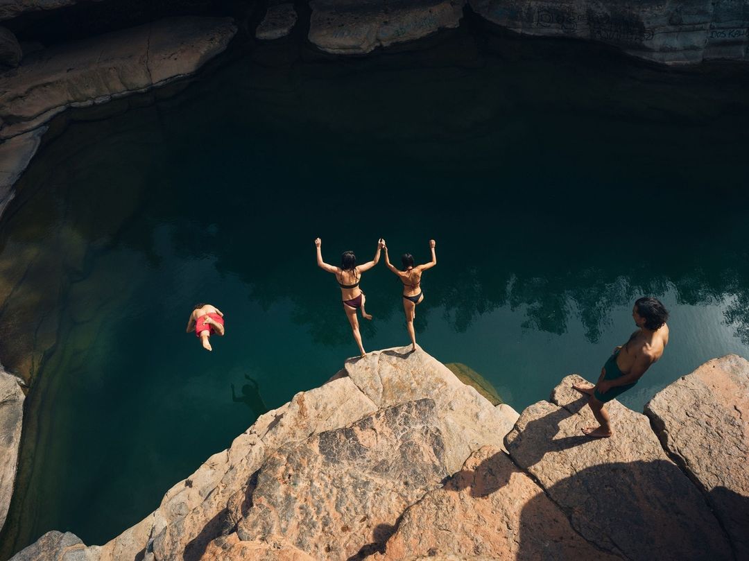 Overhead view of two women holding hands on a cliff edge, preparing to jump into a deep pool, while another person dives in.