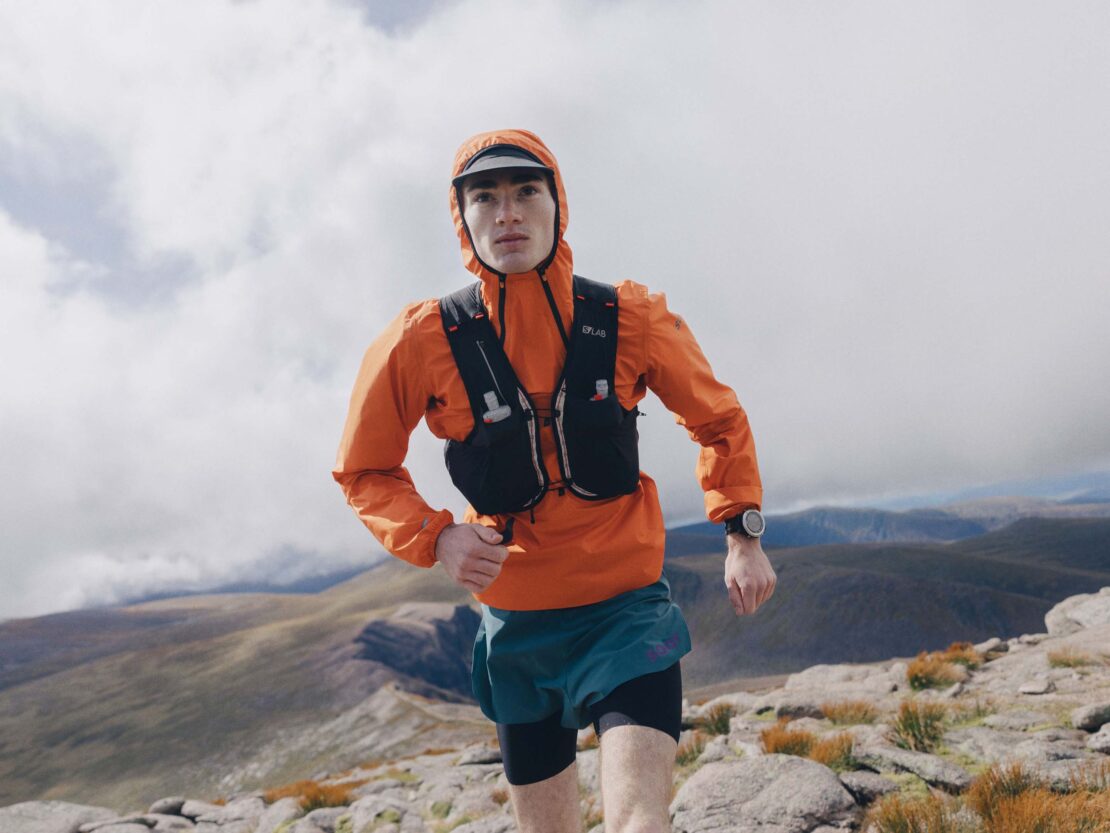 Athletic adult man running in an orange hooded jacket with mountains and cloudy sky in the background, captured in an eye-level shot.