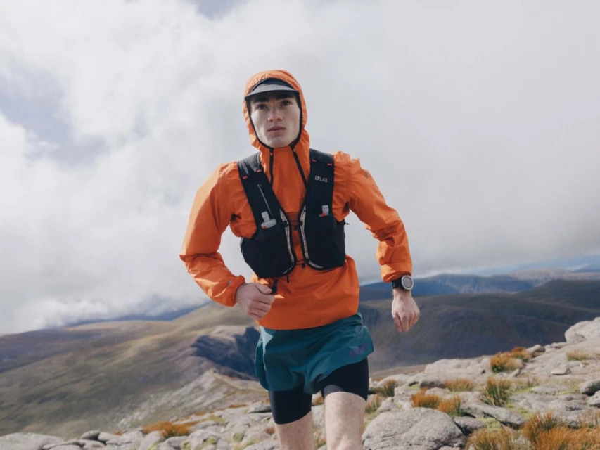 Athletic adult man running in an orange hooded jacket with mountains and cloudy sky in the background, captured in an eye-level shot.