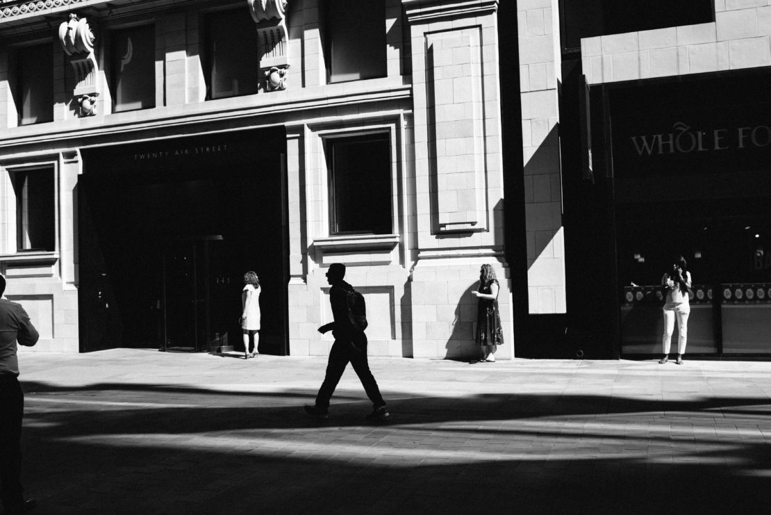 Black and white street scene featuring a man walking in the foreground, framed by architectural details and shadows, and passersby.