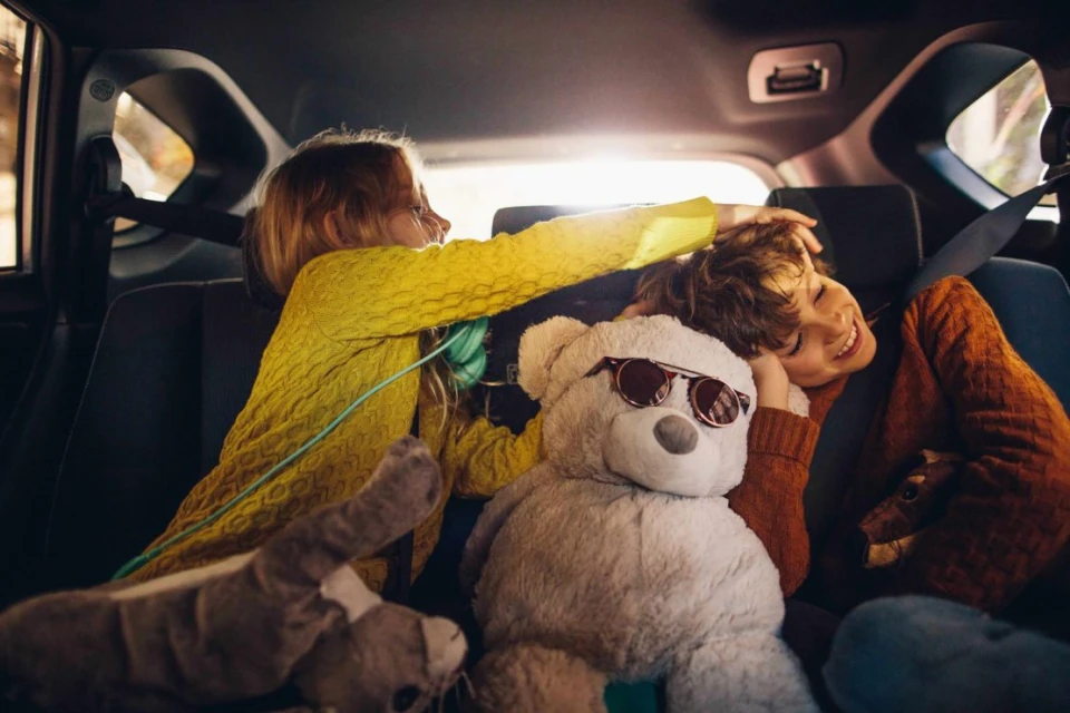 Two children, a girl and boy, ride in the back seat of a car surrounded by stuffed animals. The boy is smiling and the girl is touching his hair.