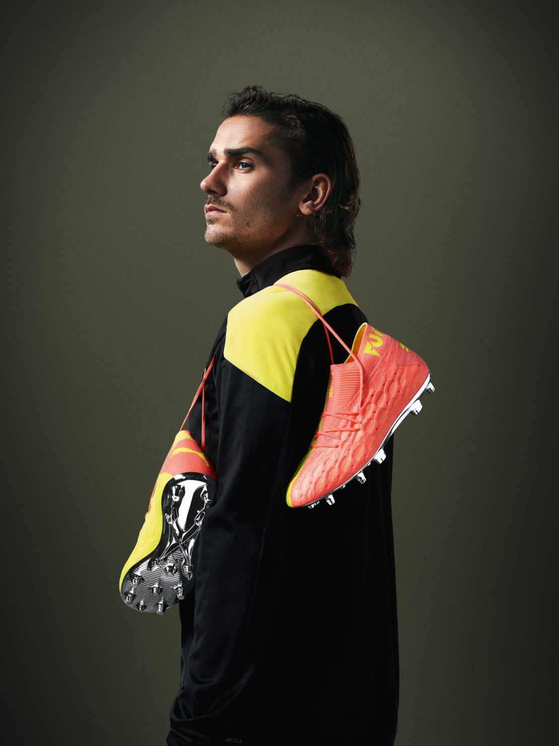 A man with dark hair poses indoors wearing a black and yellow tracksuit, with bright orange and yellow soccer shoes draped over his shoulders.