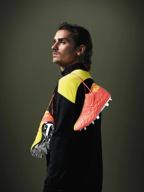 A man with dark hair poses indoors wearing a black and yellow tracksuit, with bright orange and yellow soccer shoes draped over his shoulders.