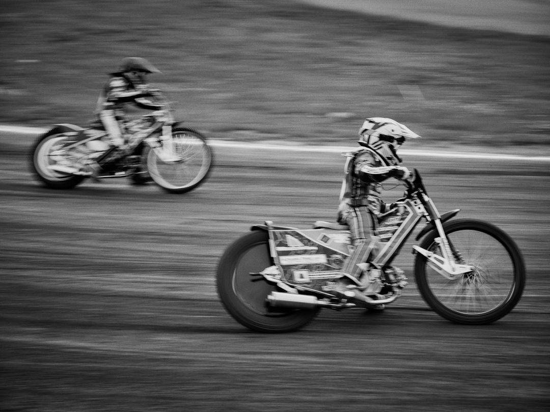 Two people speedway racing on motorcycles on a dirt track with motion blur showing speed and action in black and white.