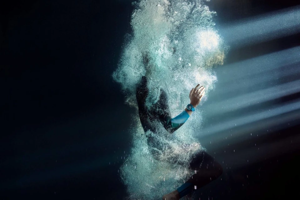 Person in wetsuit diving into dark water creating bubbles with sunlight filtering through, wearing a blue sport watch.