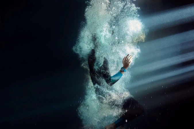 Person in wetsuit diving into dark water creating bubbles with sunlight filtering through, wearing a blue sport watch.