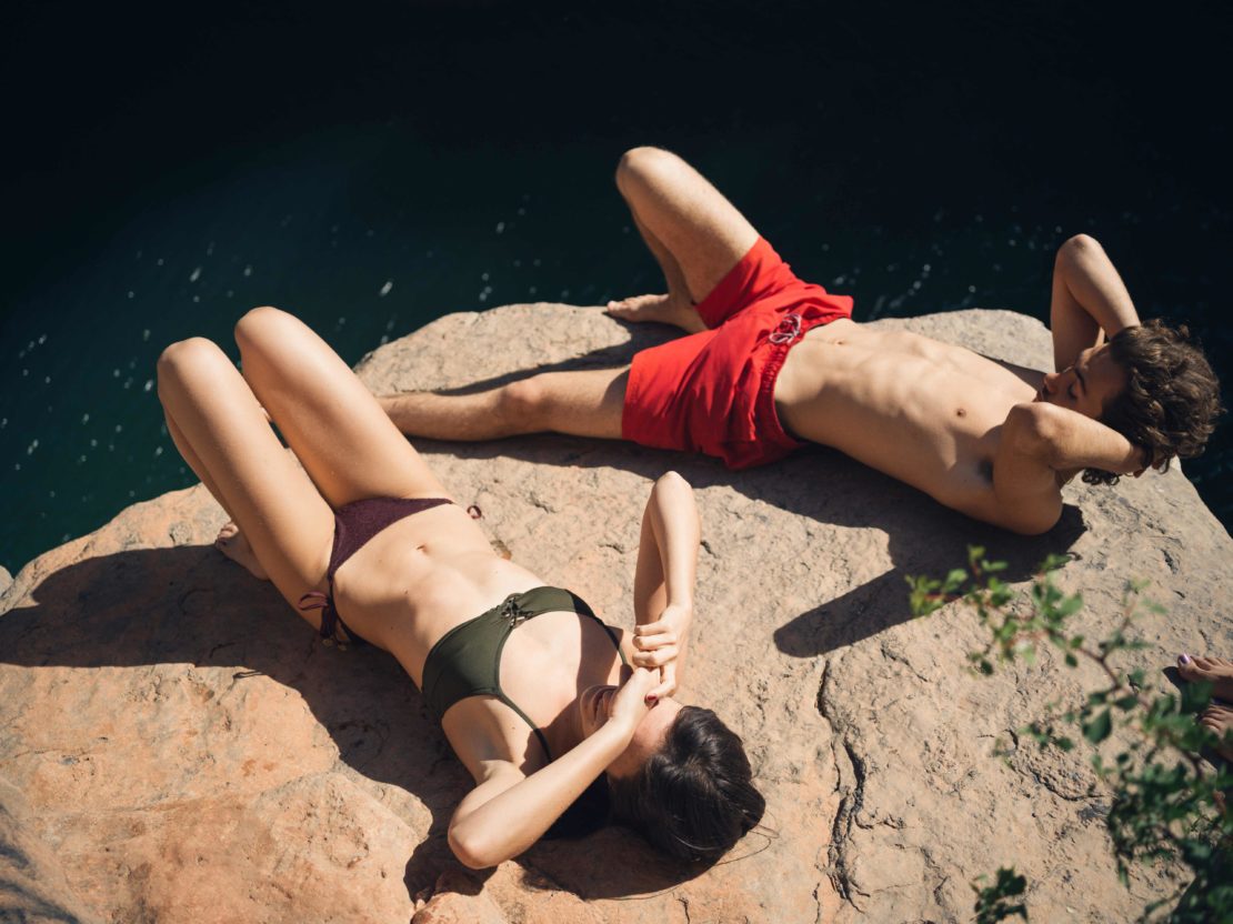Two adults, one male and one female, relaxing on a rock ledge next to dark water wearing swimwear in outdoor daylight.