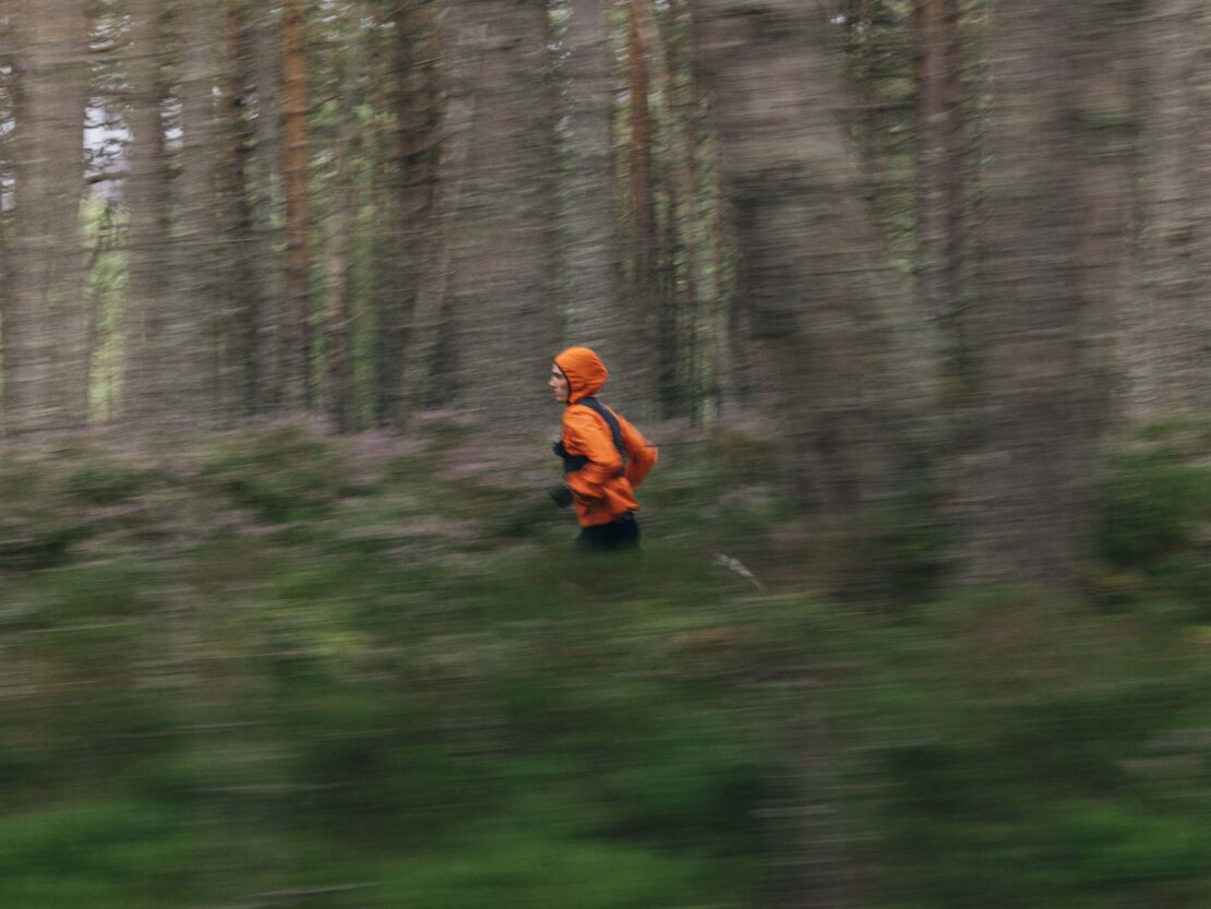 A person in an orange jacket runs through a forest with tall trees, captured with motion blur to emphasize the speed and movement.