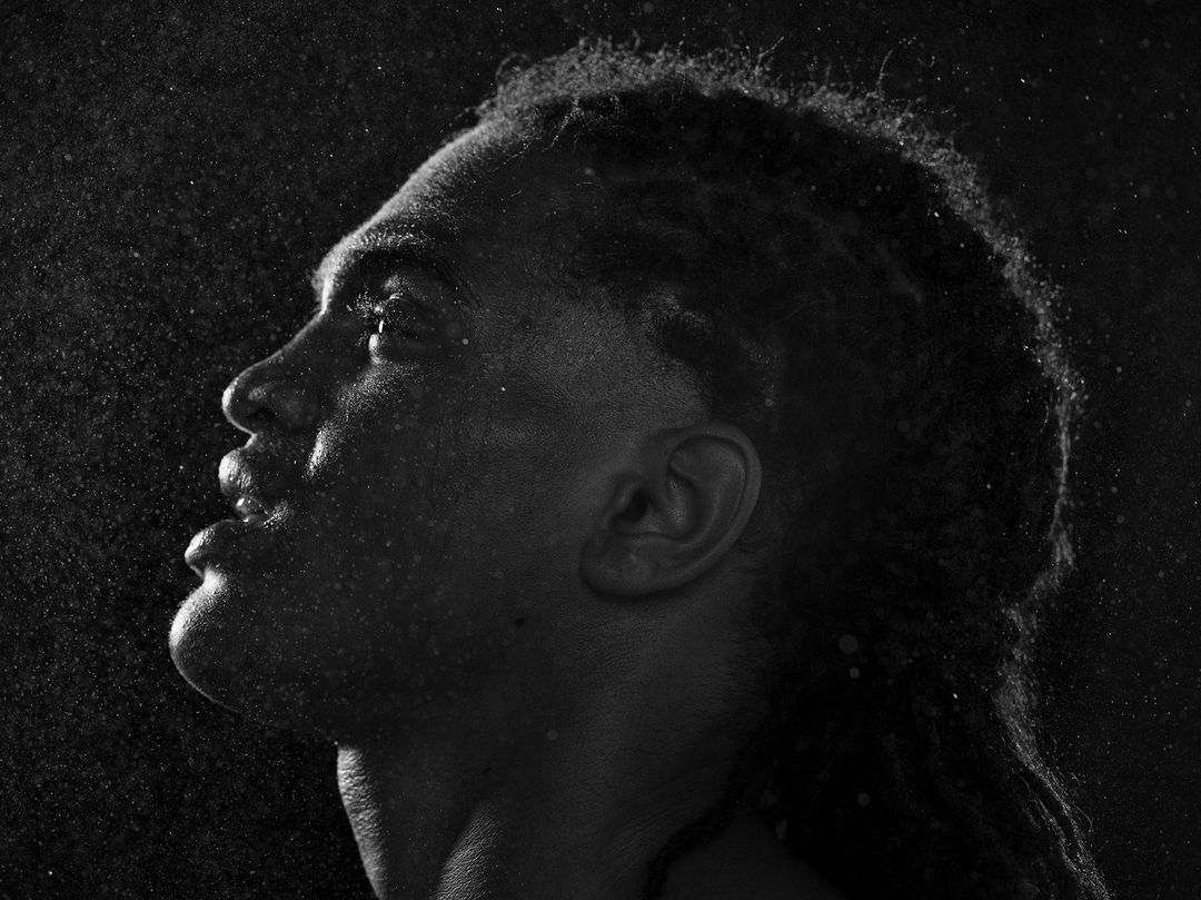 Monochrome portrait of a Black adult male with dreadlocks, looking upward, face dampened, against a dark backdrop with specks of light.