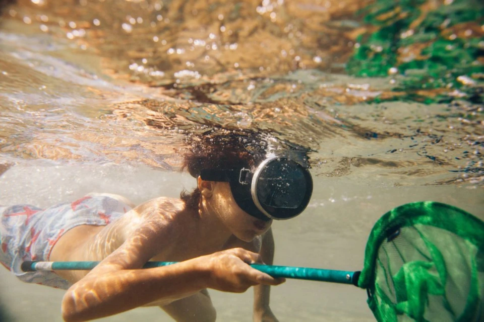 Boy with diving mask swims underwater with a green net, sunlit clear water, and sandy bottom create a natural aquatic scene.