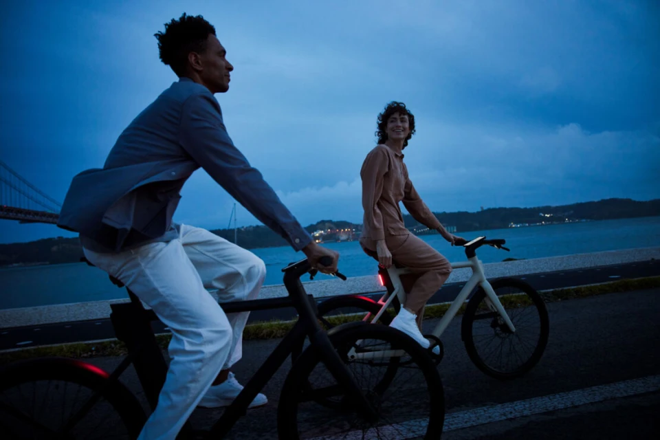 Two cyclists, an adult male and female, ride e-bikes along the coast at dusk; bridge and horizon in the background.