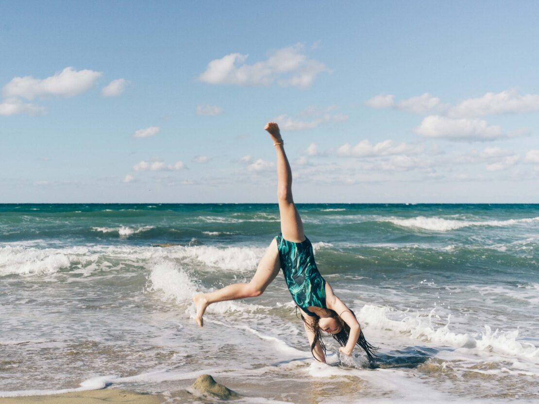 Young woman doing a cartwheel on a sandy beach near the ocean with waves under a blue sky with clouds. The ocean is turquoise.