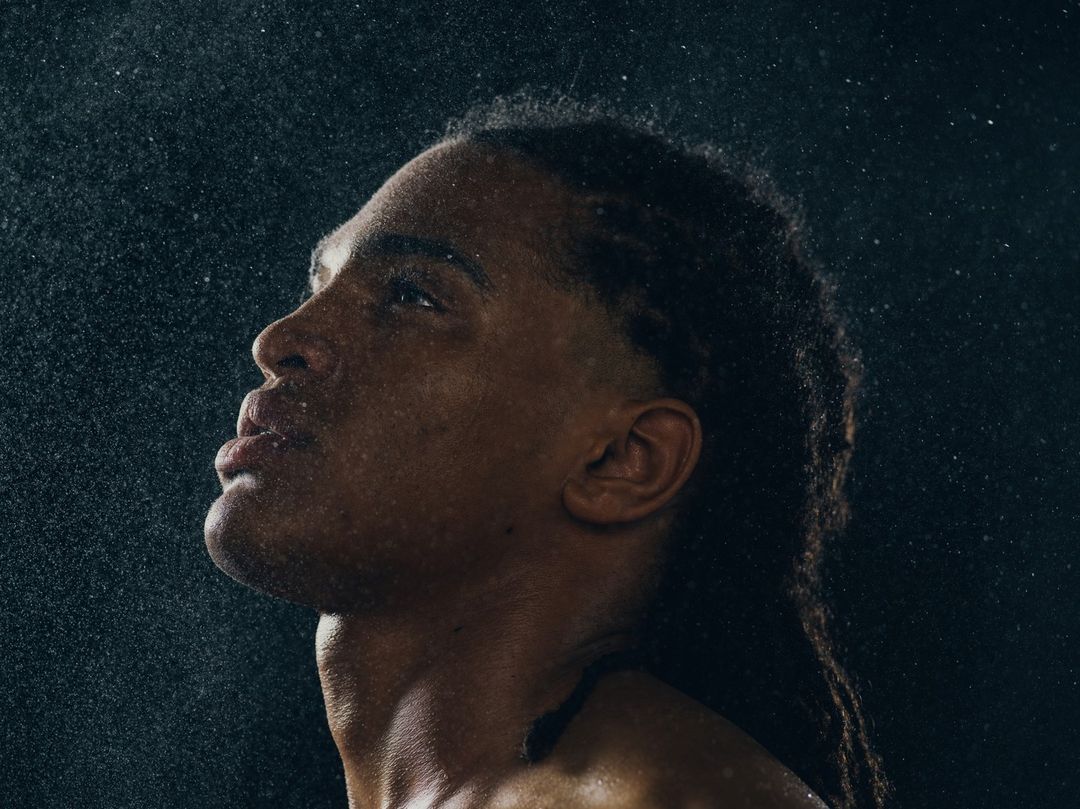 Profile shot of an adult male with dark skin and long dreadlocks, looking up while water droplets fall around him against a dark backdrop.